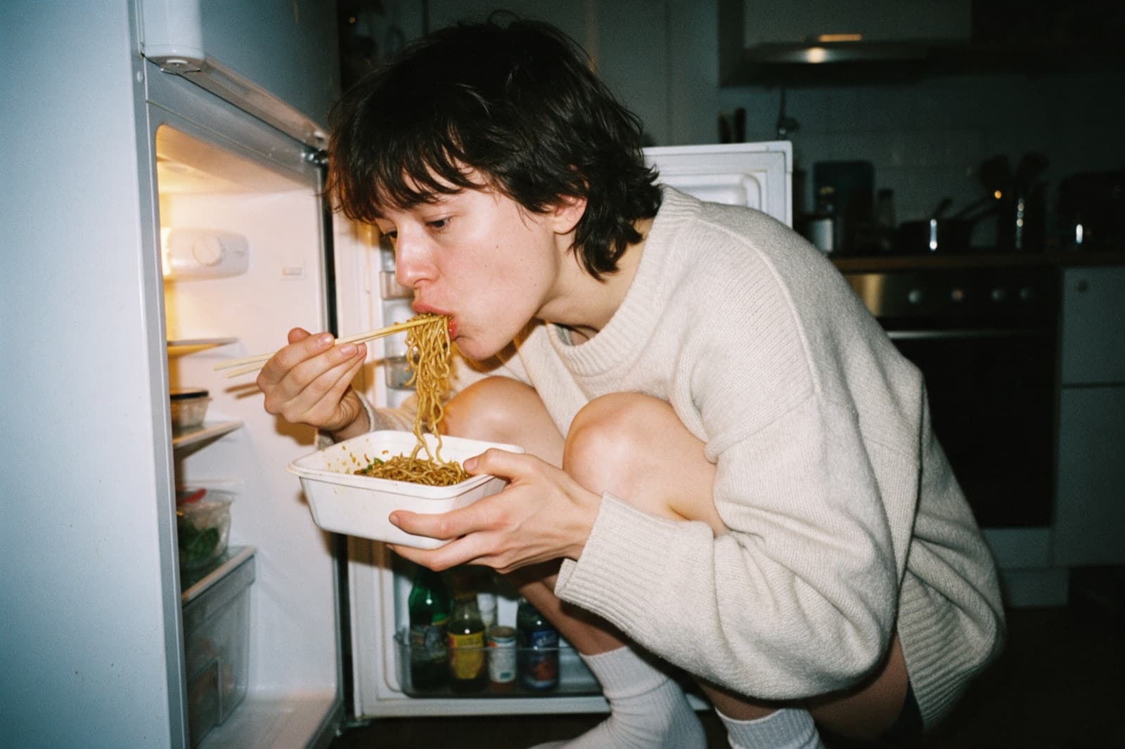 Someone crouched at an open fridge late at night, eating cold noodles from a takeaway box