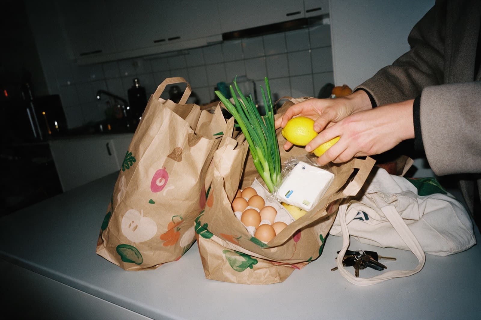 Hands unpacking lemons, spring onions, eggs and feta from paper grocery bags on a kitchen counter at night