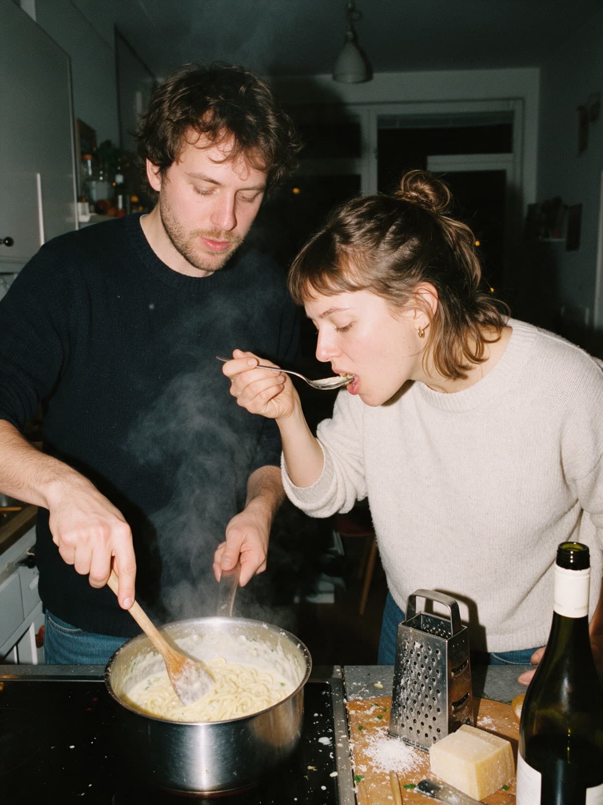 Two friends at the stove at night, one stirring a pot of creamy pasta, the other tasting from a spoon
