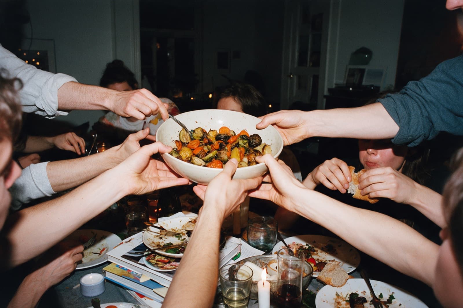 Hands passing a bowl of roast vegetables across a crowded dinner table at night